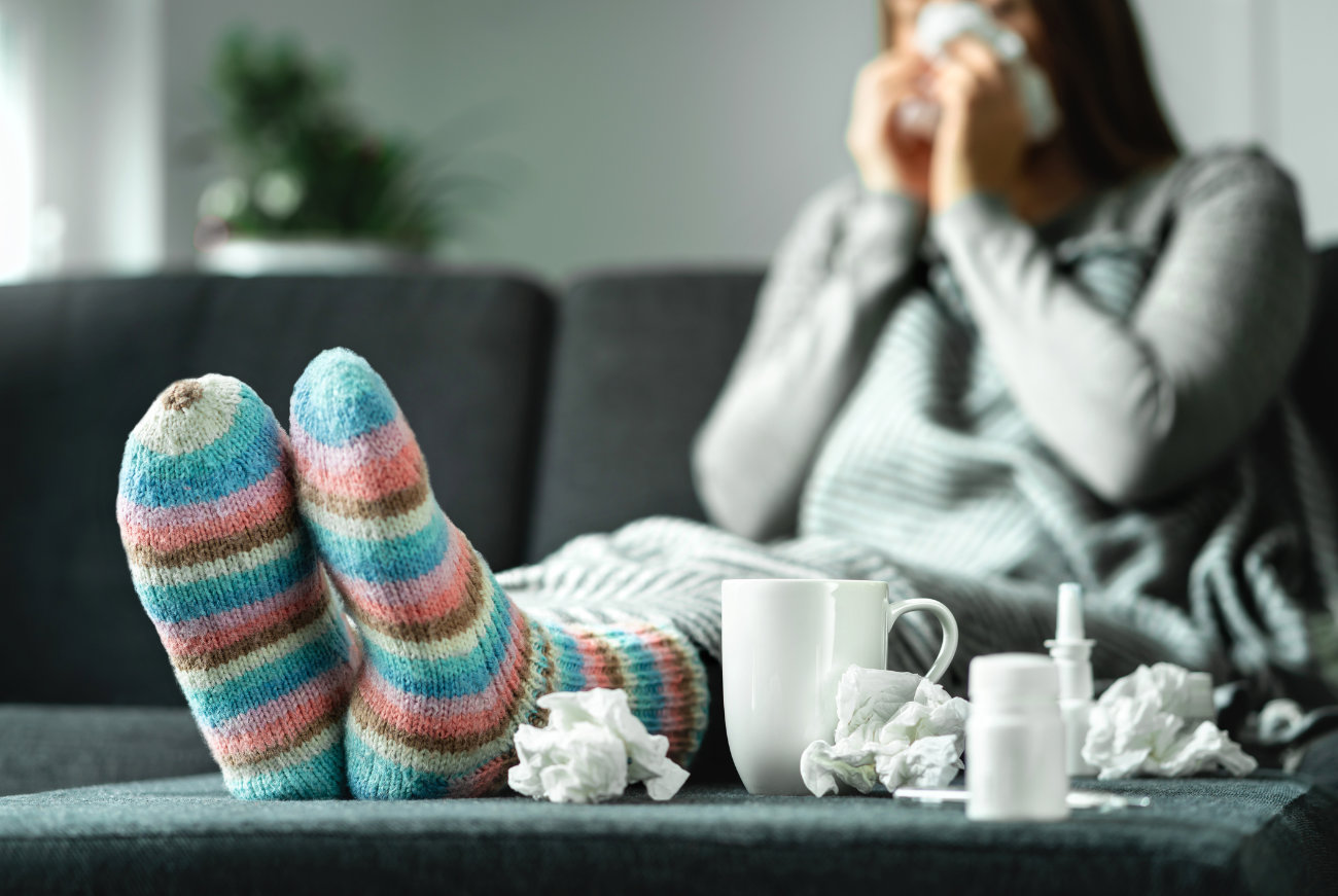 Krankheiten: Eine Frau mit einer Erkältung sitzt auf der Couch und schnäuzt sich die Nase. Sie trägt dicke, bunte Wollsocken. Vor ihr stehen eine Tasse Tee, Nasentropfen und Taschentücher.
