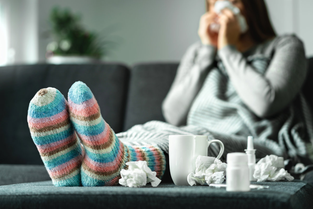 Eine kranke Frau liegt auf der Couch. Neben ihr befinden sich viele Taschentücher, eine Teetasse und ein Nasenspray