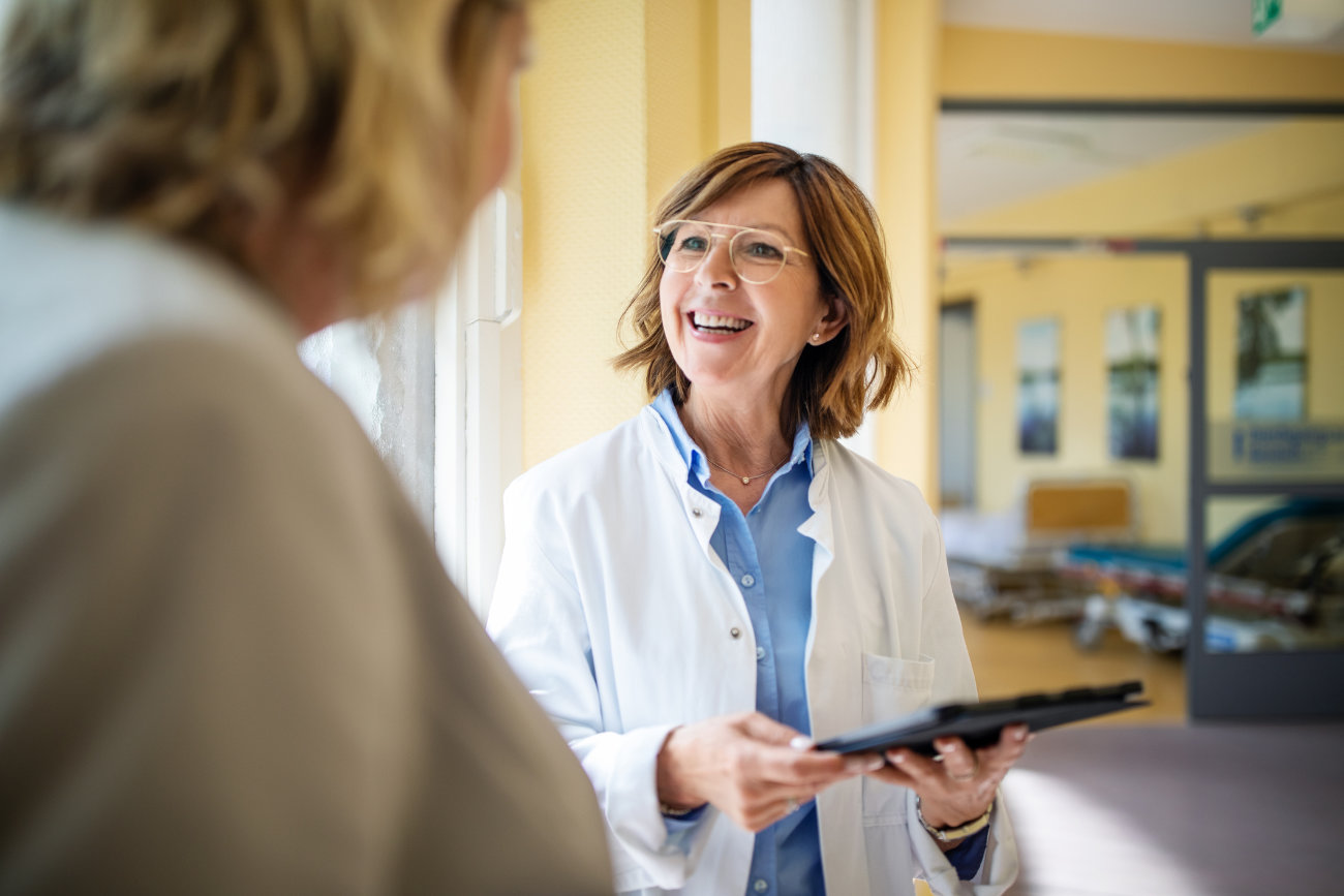 Gesundheitsversorgung Steiermark: Eine Ärztin im weißen Mantel mit schulterlangem braunen Haar und Brille steht vor einer Patientin die man nur von hinten sieht.
