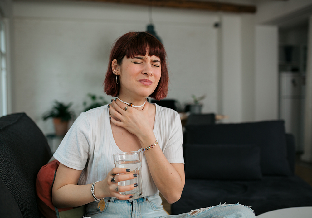 Mein Beitrag für meine Gesundheit: Ein junges Mädchen sitzt auf der Couch und greift sich auf den Hals. Ihr Gesicht ist schmerzverzerrt. Sie trägt ein weißes T-Shirt und eine Jeanshose. Die Haare sind rötlich und sie hat einen Pony.