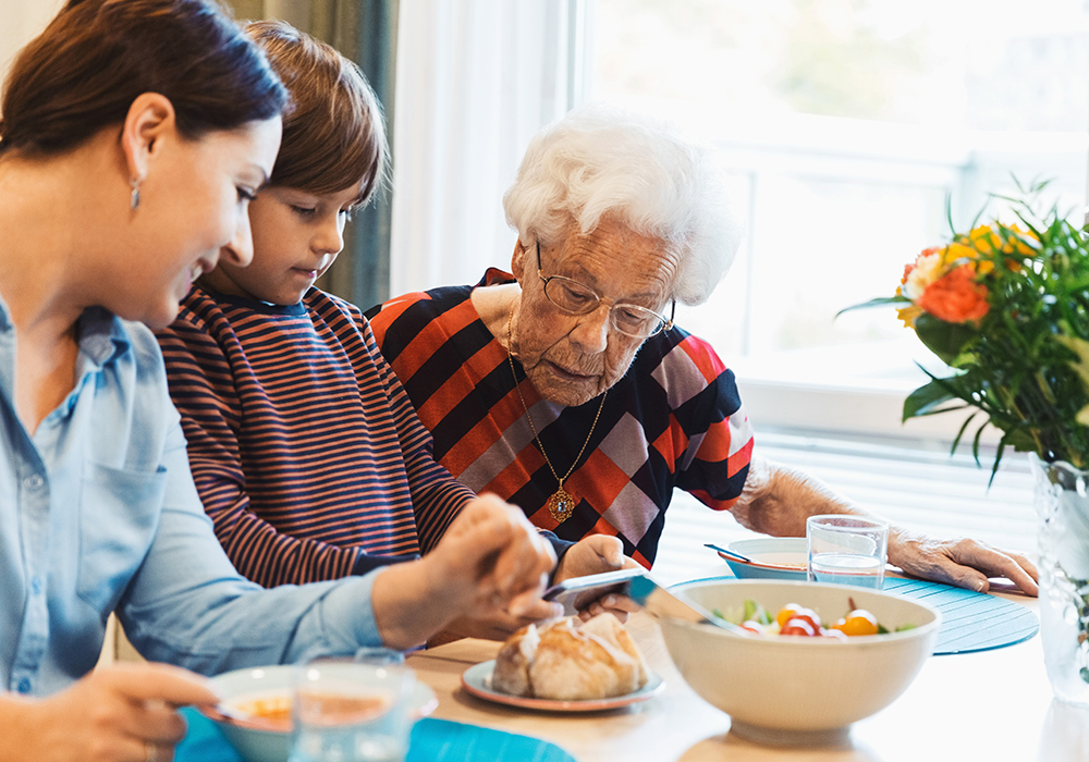 Notfall: Eine ältere Frau sitzt mit ihrer Tochter und ihrem Enkel am Tisch beim Frühstück.  Sie hat weiße, kurze Haare und eine Brille. Der Enkel zeigt ihr etwas auf dem Handy.