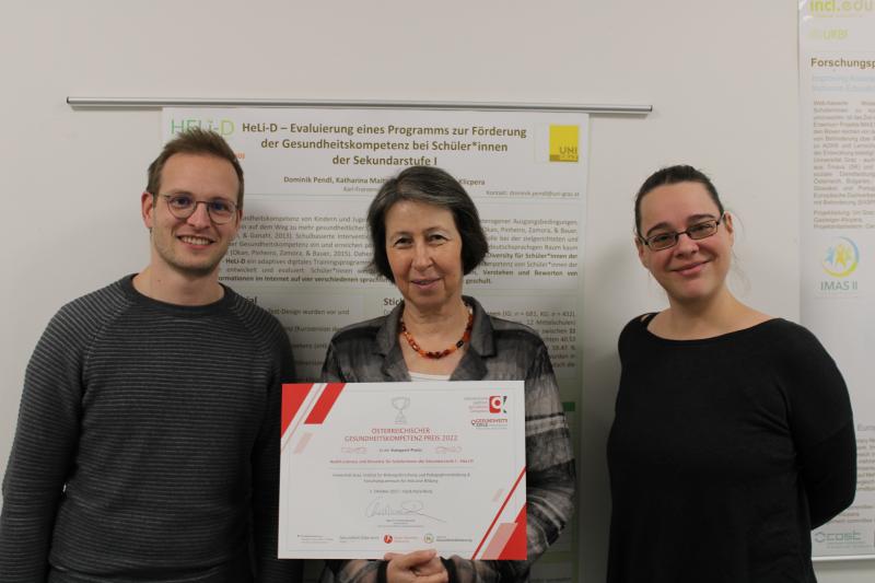 Drei Personen stehen nebeneinander, während die Frau in der Mitte eine Auszeichnung von der österreichischen Plattform für Gesundheitskompetenz in den Händen hält. 