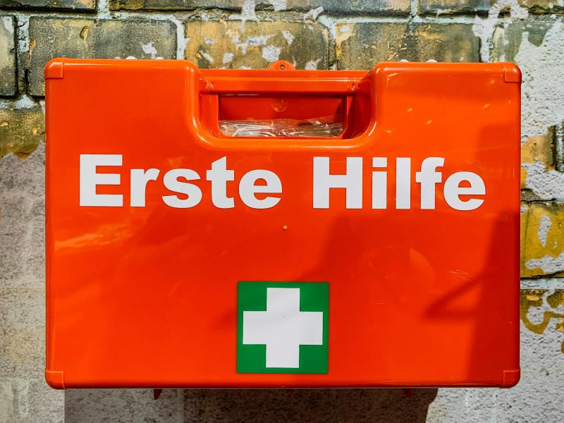 Das Bild zeigt einen orangen Erste-Hilfe-Koffer, der an einer Wand hängt. 