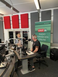 Aufnahme eines Podcasts in einem Podcast Studio. Im Hintergrund steht ein Banner mit der Aufschrift ORF St Radio, Fernsehen, Online