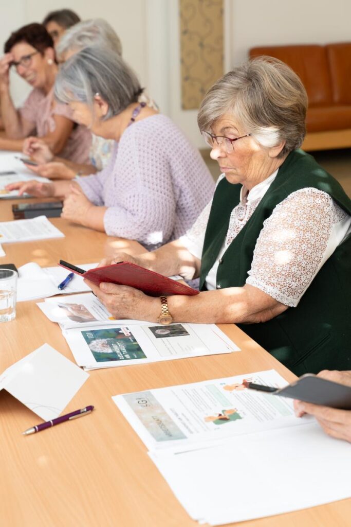 Seniorinnen sitzen gemeinsam an einem Tisch und beschäftigen sich mit Tablets und gedruckten Unterlagen