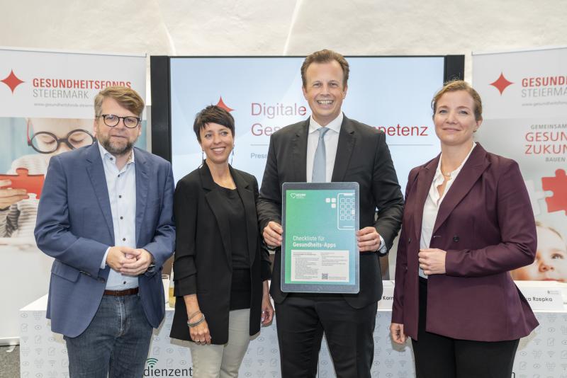 Fünf Personen stehen nebeneinander und lächeln in die Kamera, eine Person in der Mitte hält eine Auszeichnung mit der Aufschrift ‚Checkliste für Gesundheits-Apps‘. Im Hintergrund sind Roll-Ups mit dem Logo des Gesundheitsfonds Steiermark zu sehen