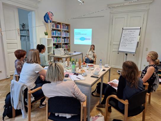 Eine Lerngruppe, die um einen Tisch sitzt und etwas von einer Person aus der Gruppe erklärt bekommt. Es ist außerdem ein Flipchart und eine Powerpointpräsentation in dem Bild zu erkennen.