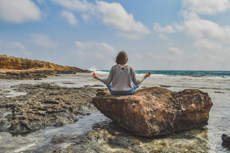 Frau sitzt im Schneidersitze auf einem Fels und schaut aufs Meer.