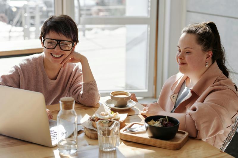 Zwei Frauen sitzen an einem Tisch in einem Café, unterhalten sich und genießen eine Mahlzeit; vor ihnen stehen ein Laptop, Kaffee, Wasser und Speisen.