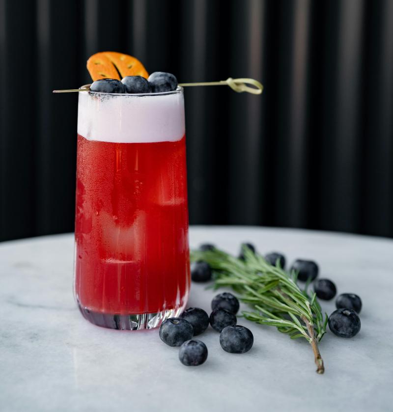 Roter Cocktail mit Schaumhaube in einem hohen Glas, garniert mit Heidelbeeren und Orangenscheibe auf einem Holzspieß; daneben frische Heidelbeeren und Rosmarinzweig auf Marmortisch