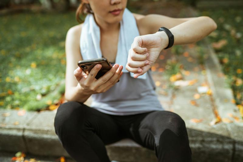 Junge Frau im Sportoutfit sitzt draußen auf einer Bank und nutzt eine Gesundheits-App auf Smartphone und Smartwatch