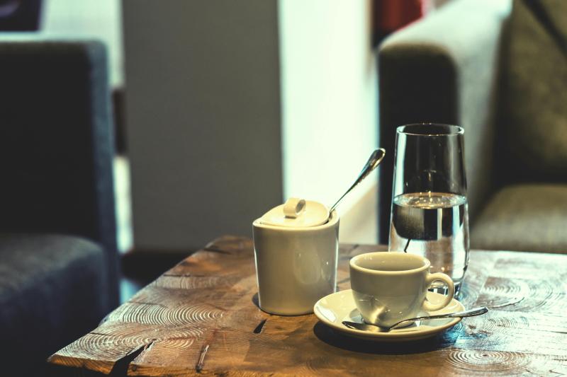 Holztisch mit einer Tasse Kaffee auf Untertasse, einem Glas Wasser und einem weißen Zuckerdöschen mit Löffel, im Hintergrund gemütliche Sessel.