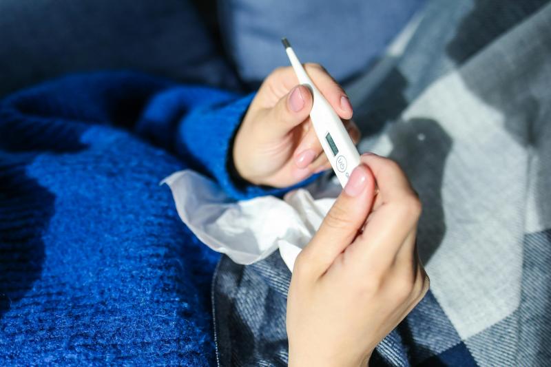 Person hält ein digitales Fieberthermometer und ein Taschentuch, liegt eingehüllt in eine blaue Decke